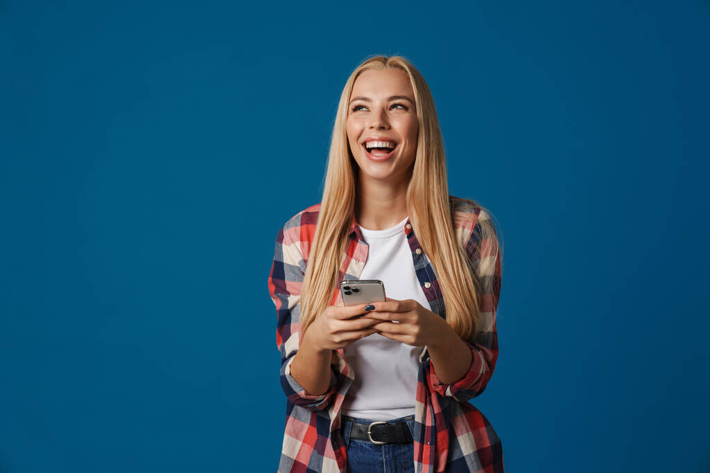 Blondynka szczęśliwa biała kobieta śmieje się podczas korzystania z telefonu komórkowego odizolowany na niebieskim tle - Zdjęcie, obraz