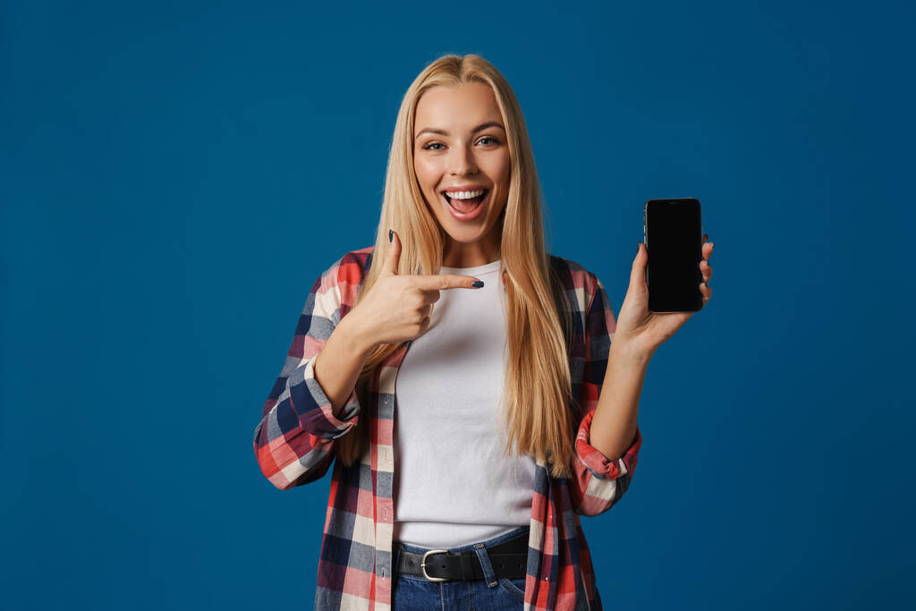 Весела блондинка показує і вказує палець на смартфон ізольовано на синьому фоні
 - Фото, зображення