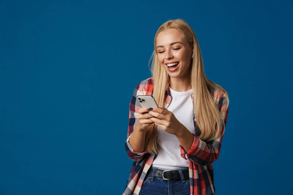 Veselé krásné blondýny dívka smějící se a pomocí smartphone izolované přes modré pozadí - Fotografie, Obrázek