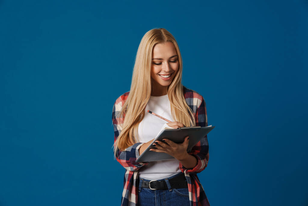 Happy beautiful blonde girl smiling while writing down notes isolated over blue background - 写真・画像