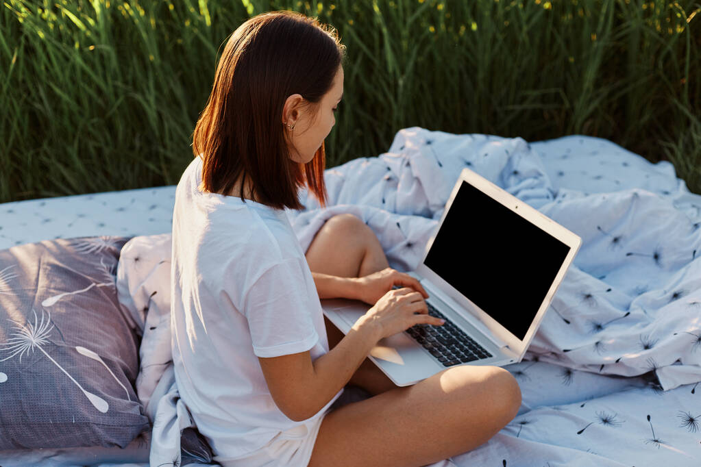 黒い髪の女性のサイドビュー白いカジュアルなTシャツを身に着けている横断脚と空の画面でトップラップを保持し、フリーランスは牧草地で柔らかいベッドの上でオンラインで作業. - 写真・画像
