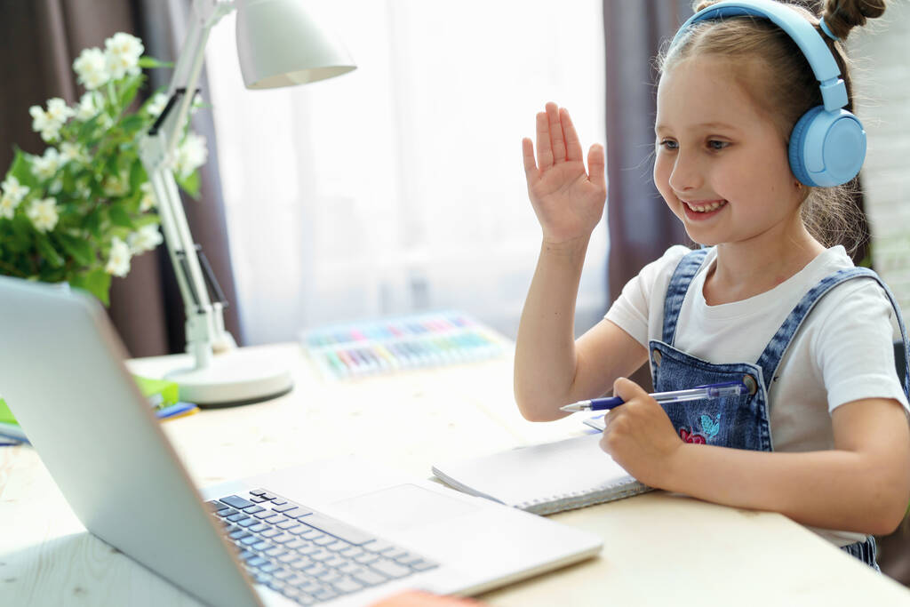 Kulaklıklı Shcoolgirl karantina döneminde uzaktan öğrenmek için dizüstü bilgisayar kullanıyor. Çocuk derse cevap vermek için elini kaldırır. E-eğitim. Uzaklık Öğrenme, Evde Öğrenme. - Fotoğraf, Görsel