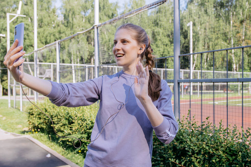 Egy sportos egyenruhás lány az edzőteremben fülhallgatóban és egy telefon a kezében üdvözli a beszélgetőtársát a jobb kezével. - Fotó, kép