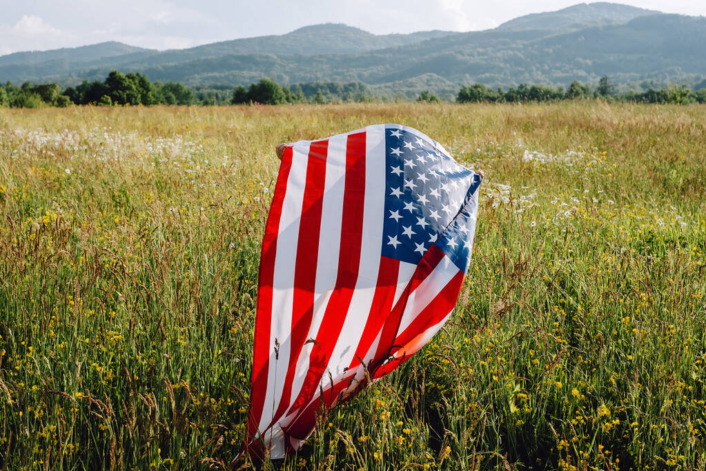 Pohled zezadu nepoznatelné malé holčička stojící na zelené trávě s vlajkou Spojených států těší západ slunce na přírodě. 4. července, oslava Dne nezávislosti. Vlastenecká dovolená, koncept demokracie - Fotografie, Obrázek