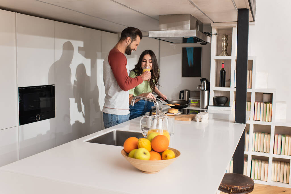 Mosolygós férfi kezében pohár narancslé közelében barátnője serpenyő a konyhában  - Fotó, kép
