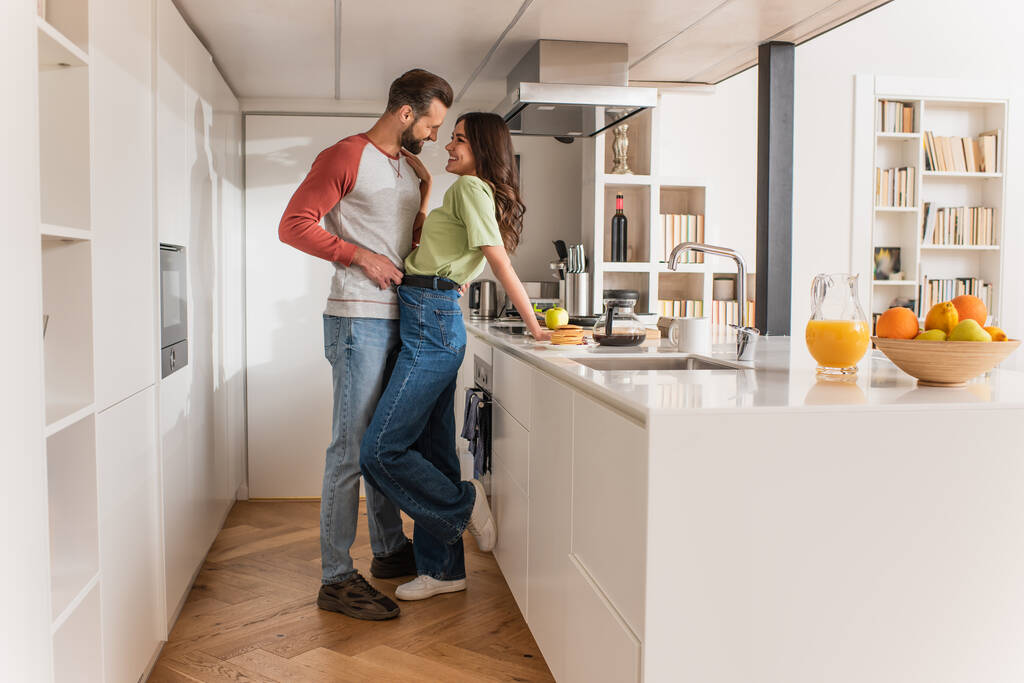 Πλευρική άποψη του ανθρώπου αγκαλιάζει χαμογελαστή φίλη κοντά στο πρωινό και τα ποτά στην κουζίνα  - Φωτογραφία, εικόνα