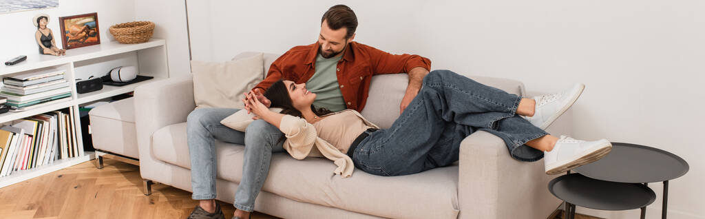 Mujer alegre cogida de la mano del novio mientras está acostado en el sofá, pancarta  - Foto, Imagen