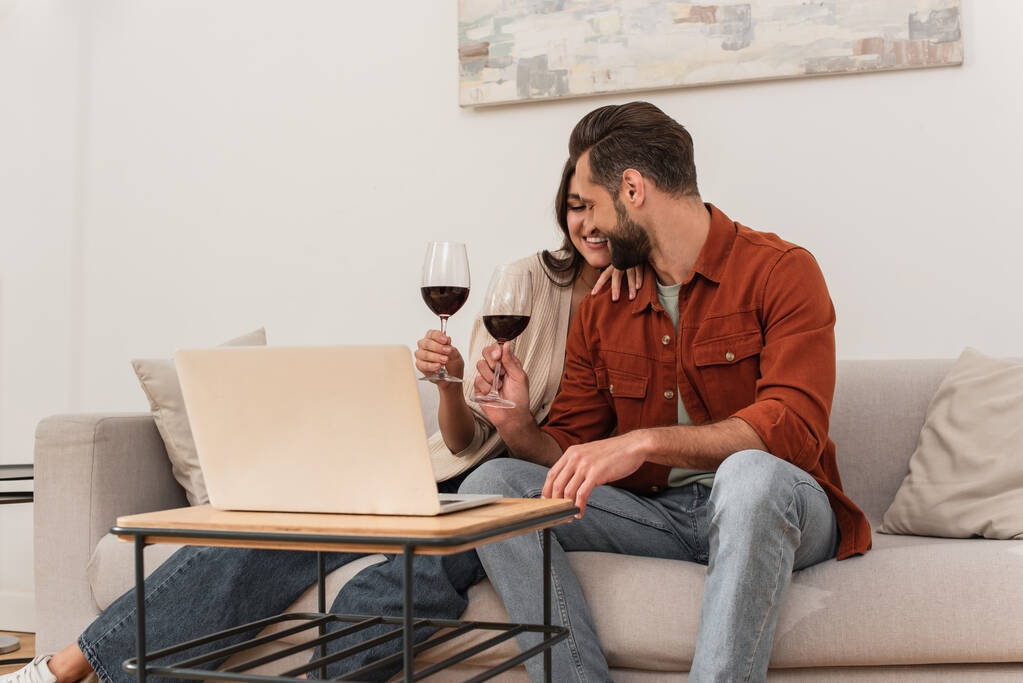 Coppia sorridente con vino seduto vicino al computer portatile in soggiorno  - Foto, immagini