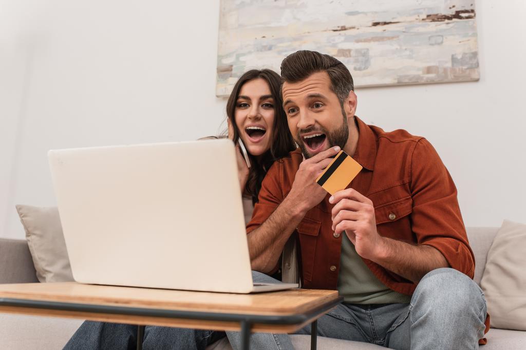Casal animado com cartão de crédito e smartphone olhando para laptop em casa  - Foto, Imagem