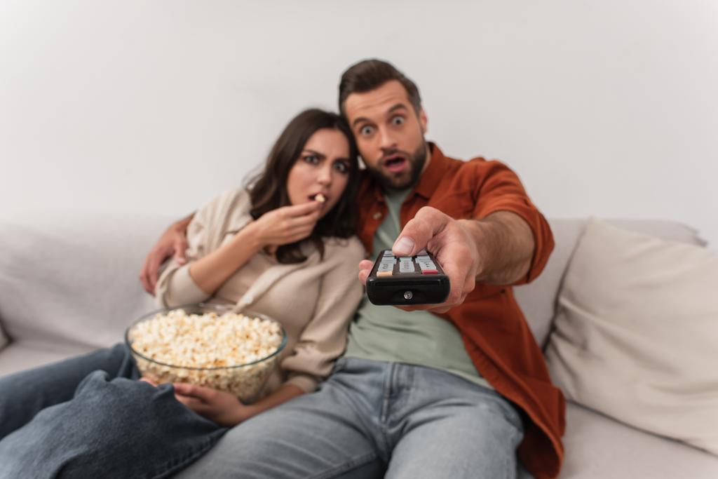 Dálkový ovladač v ruce rozmazaného muže v blízkosti užaslé přítelkyně s popcornem  - Fotografie, Obrázek