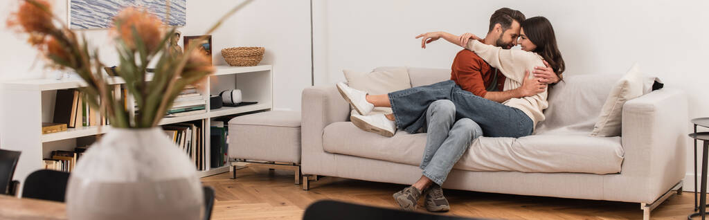 Vista lateral de la pareja joven abrazándose en el sofá en casa, pancarta  - Foto, Imagen