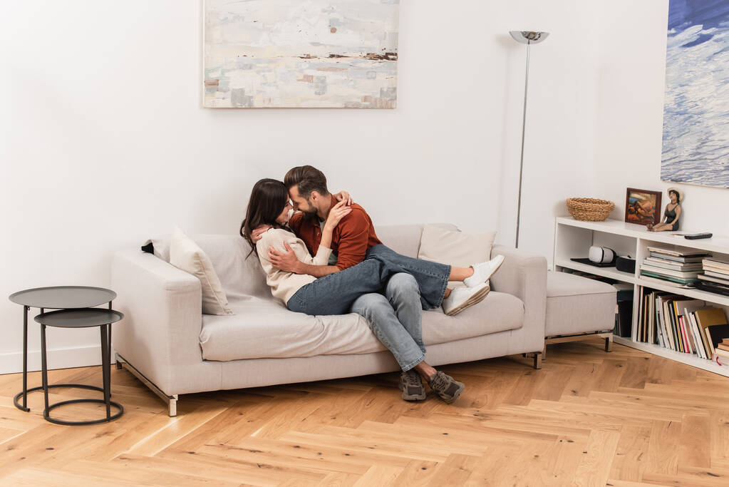 Vue latérale du couple souriant embrassant sur le canapé à la maison  - Photo, image