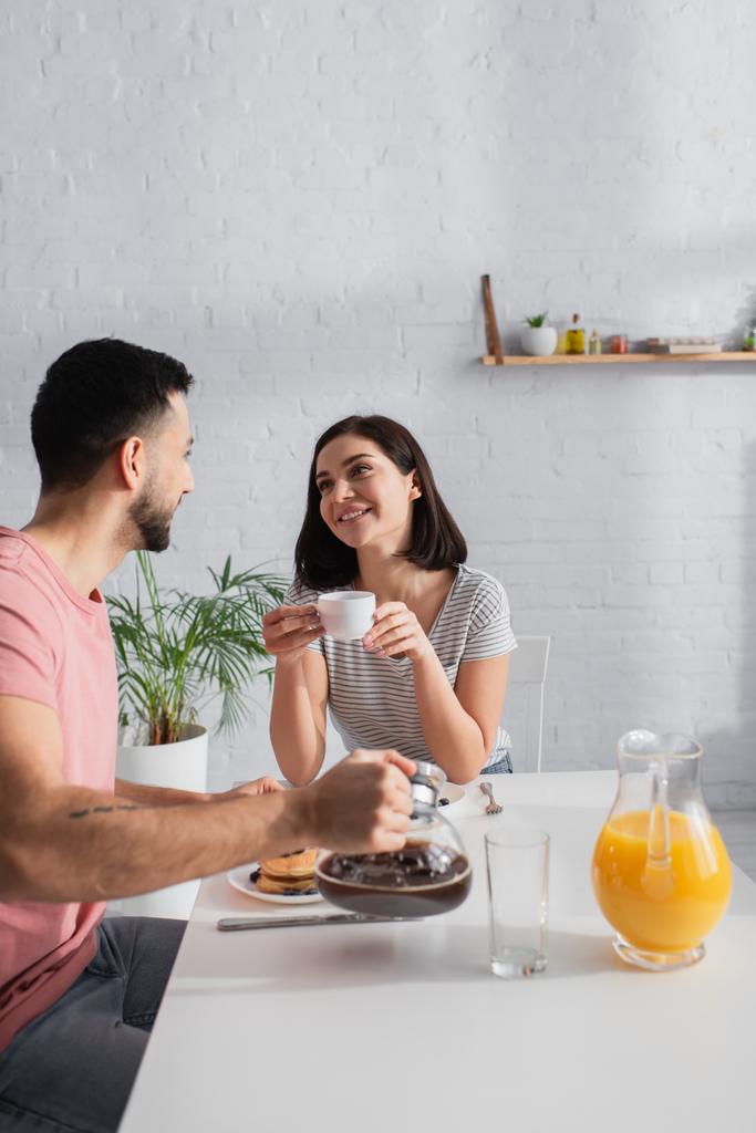 усміхнена молода жінка з чашкою кави дивиться на хлопця з кав'ярнею на кухні
 - Фото, зображення