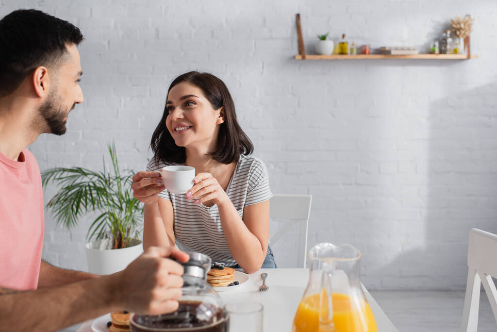 usmívající se mladá žena s šálkem v ruce při pohledu na přítele drží kávovar v kuchyni - Fotografie, Obrázek