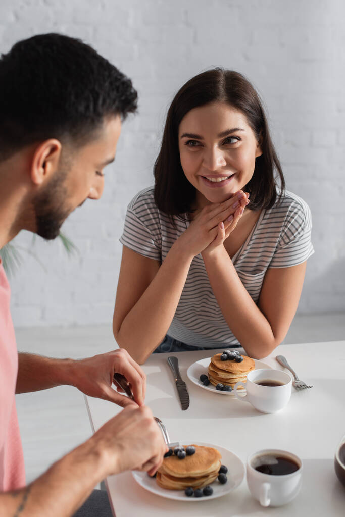 hymyilevä nuori nainen kädet lähellä kasvoja katsomalla poikaystävä syö pannukakkuja keittiössä - Valokuva, kuva