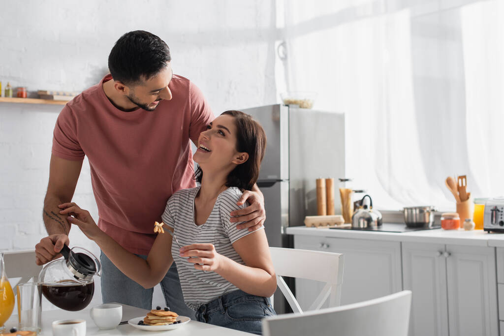 χαμογελαστός νεαρός άνδρας αγκαλιάζει τη φίλη του με κομμάτια από τηγανίτες στο πιρούνι και ρίχνει καφέ από κατσαρόλα σε φλιτζάνι στην κουζίνα - Φωτογραφία, εικόνα