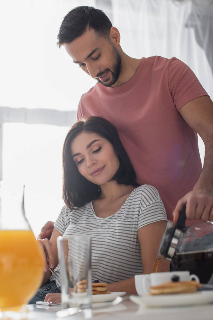 uśmiechnięty młody mężczyzna przytula dziewczynę ze śniadaniem i nalewa kawę od garnka do filiżanki w kuchni - Zdjęcie, obraz