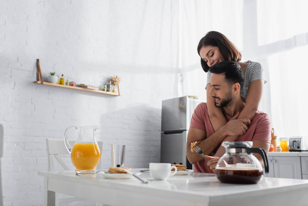giovane donna sorridente che abbraccia dolcemente il fidanzato vicino al tavolo con colazione, succo d'arancia e caffè in cucina - Foto, immagini