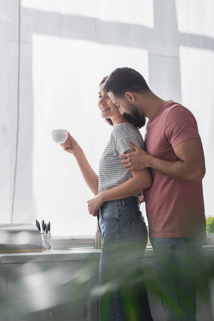 smiling young man gently kissing girlfriend with white coffee cup in modern kitchen - Photo, Image