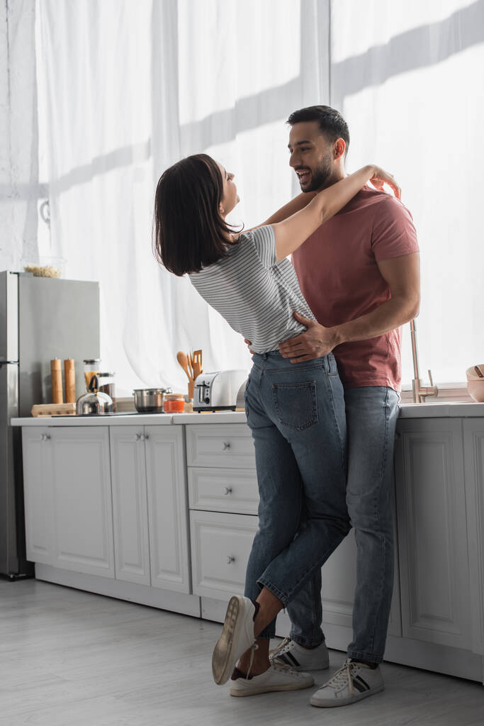positiva giovane donna abbracciare fidanzato con le mani sulle spalle in cucina - Foto, immagini