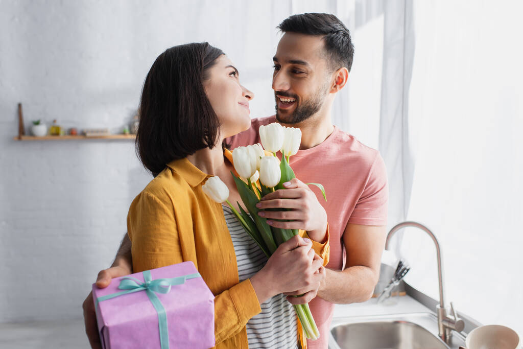 Улыбающийся молодой человек обнимает девушку и представляет букет цветов и подарочную коробку на кухне - Фото, изображение