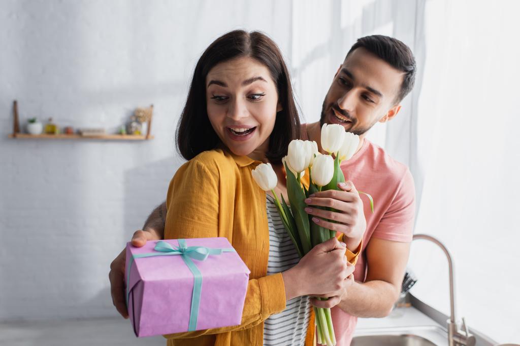 young man hugging excited girlfriend and presenting bouquet of flowers and gift box in kitchen - Photo, Image