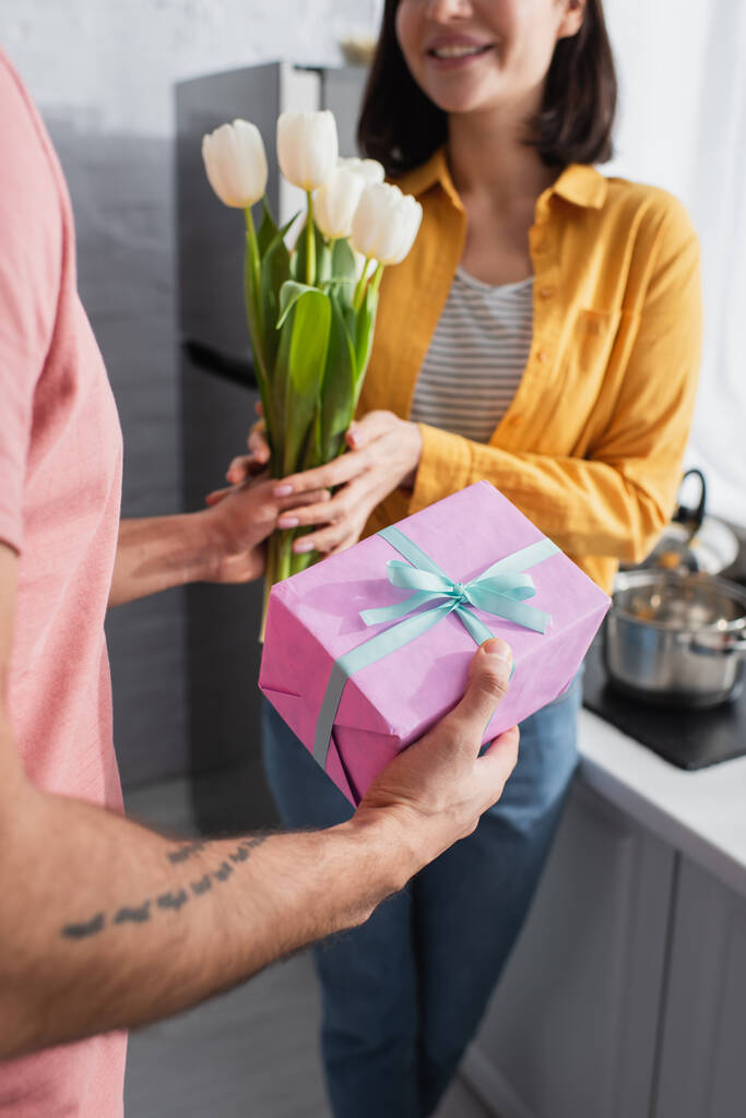 gedeeltelijk uitzicht van de jongeman presenteren boeket bloemen en geschenkdoos aan vriendin in de keuken - Foto, afbeelding