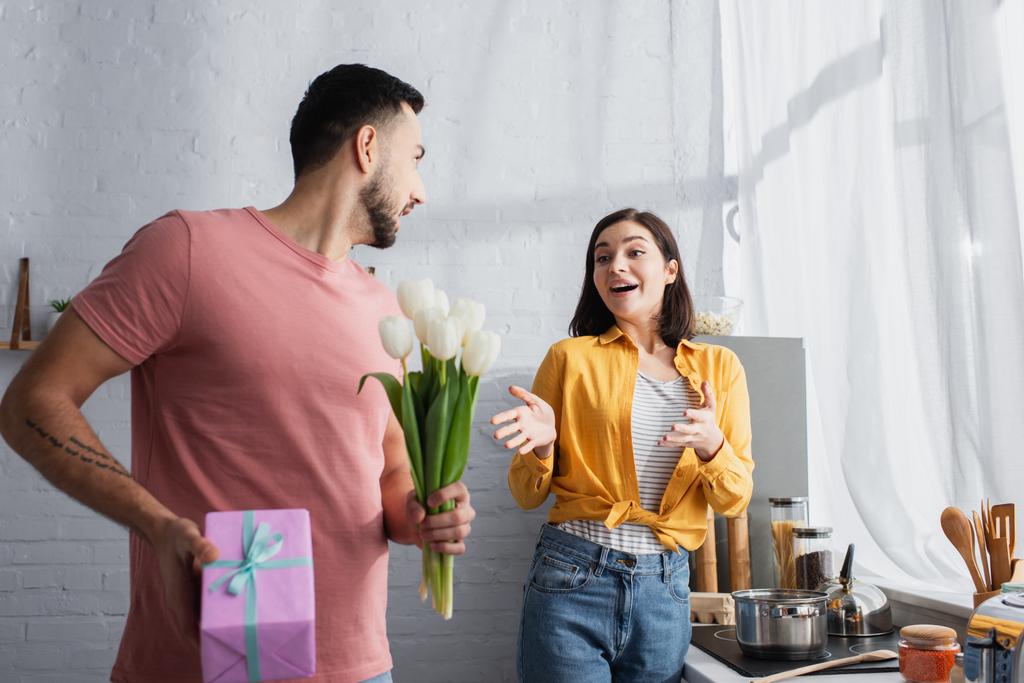 mujer joven excitada con las manos extendidas de pie cerca del hombre con ramo de flores y caja de regalo en la cocina - Foto, imagen