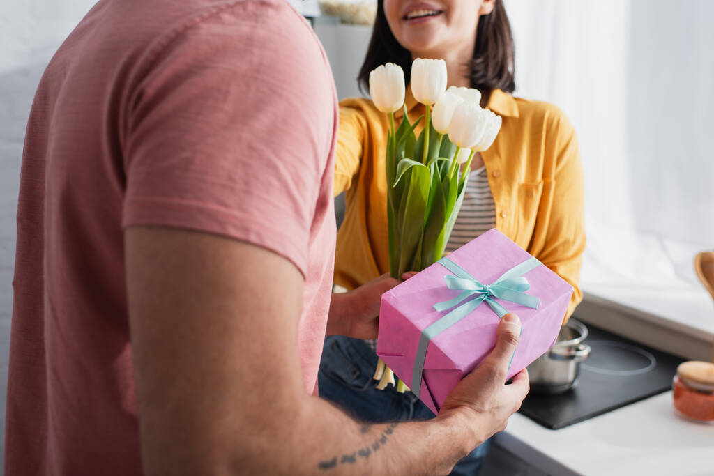 partial view of young man presenting bouquet of flowers and gift box to girlfriend in kitchen - Photo, Image