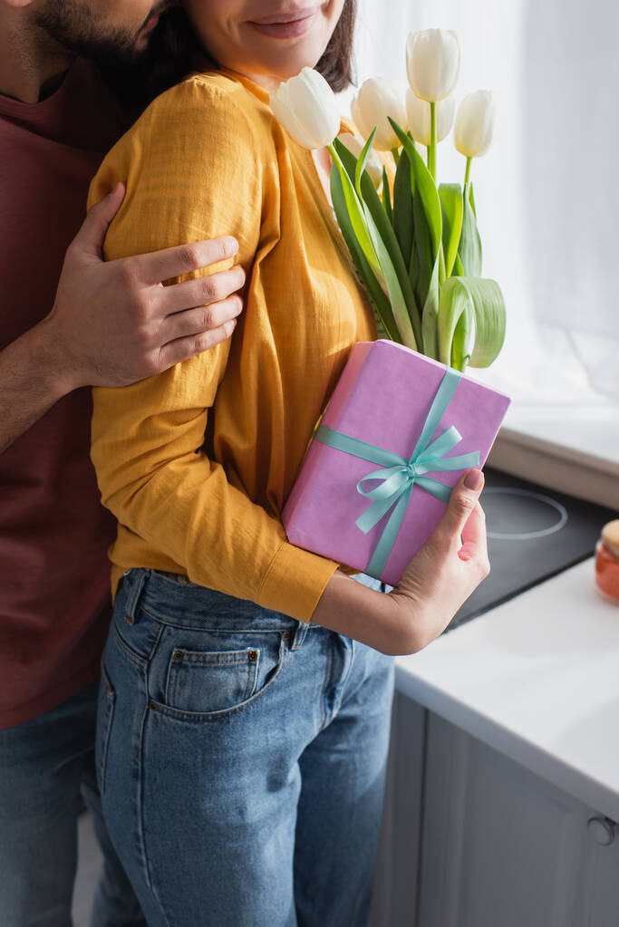 vista parcial de joven abrazando novia con ramo de flores y caja de regalo en la cocina - Foto, imagen
