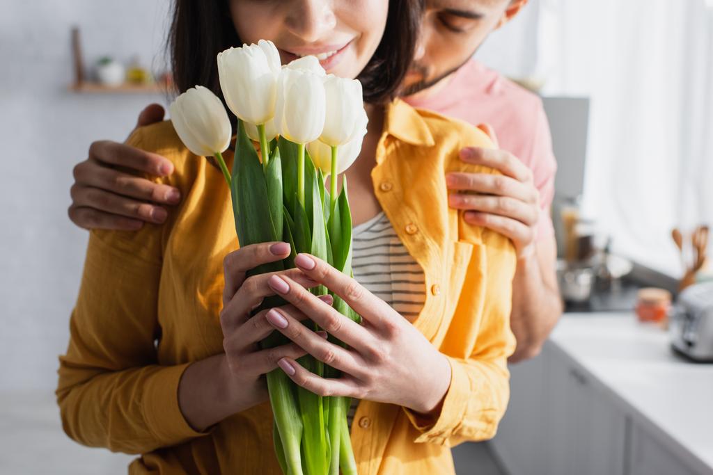částečný pohled na mladého muže objímání přítelkyně s kyticí květin v kuchyni - Fotografie, Obrázek