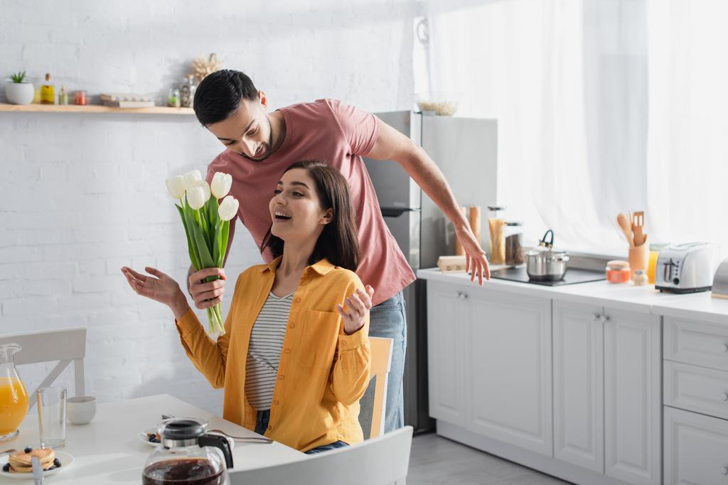 Lächelnder junger Mann überreicht aufgeregten Freundin mit offenem Mund und ausgestreckten Händen Blumenstrauß in der Küche - Foto, Bild