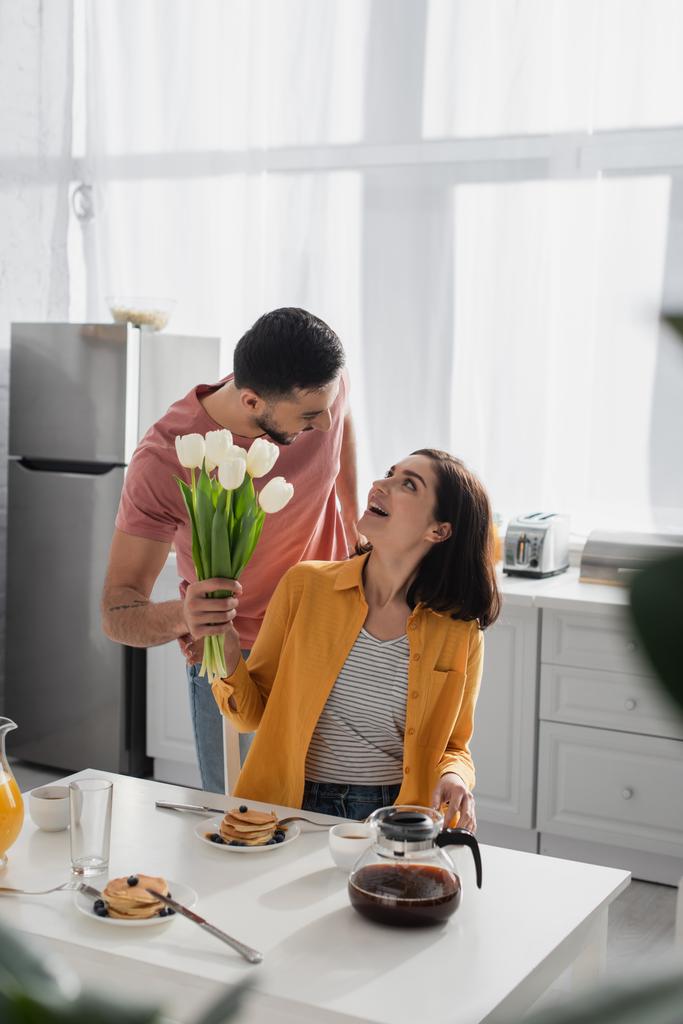 positive young man presenting bouquet of flowers to excited girlfriend with open mouth in kitchen - Photo, Image