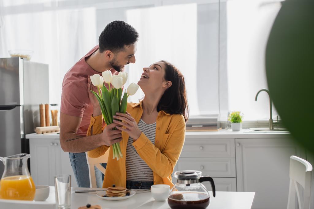 jeune homme présentant bouquet de fleurs à petite amie assise sur la table dans la cuisine - Photo, image