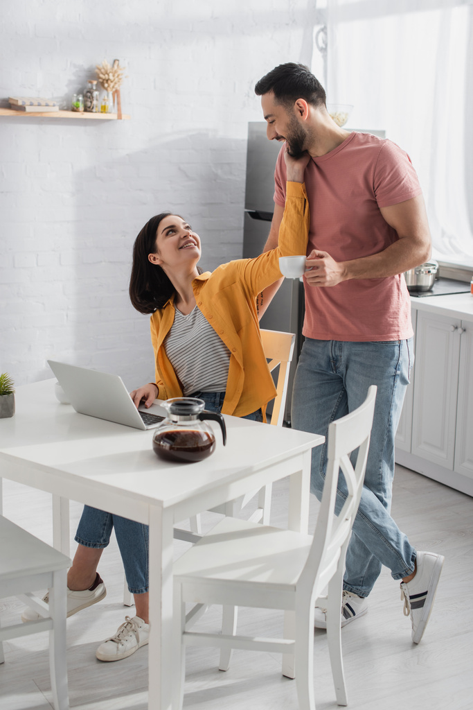sorridente giovane uomo in piedi tazza di caffè vicino fidanzata con computer portatile in cucina - Foto, immagini