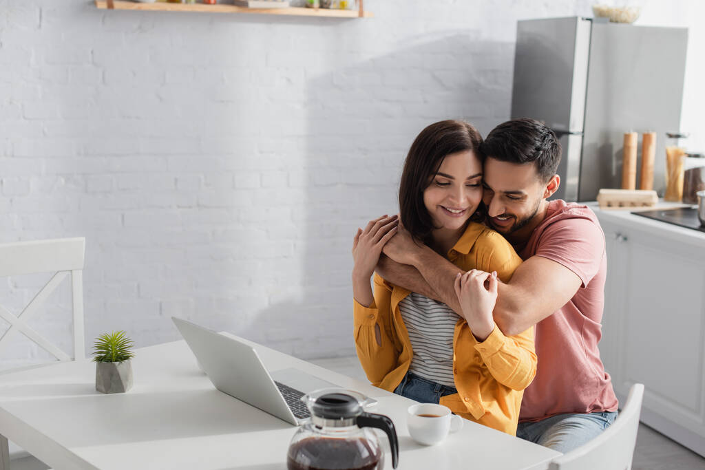 smiling young man hugging girlfriend with laptop in kitchen - Photo, Image
