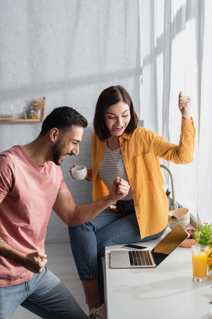 excité jeune couple regardant ordinateur portable avec des gestes de victoire à la maison - Photo, image
