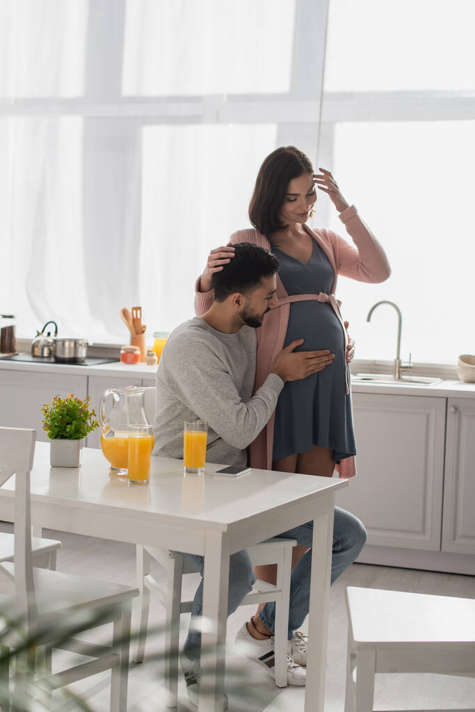 young man touching belly of smiling pregnant woman in kitchen - Photo, Image