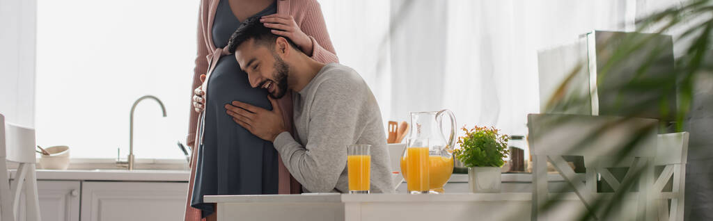 jonge man knuffelen buik van zwangere vrouw in keuken, banner - Foto, afbeelding