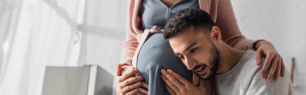 joven abrazando suavemente el vientre de la mujer embarazada en la cocina, pancarta - Foto, Imagen