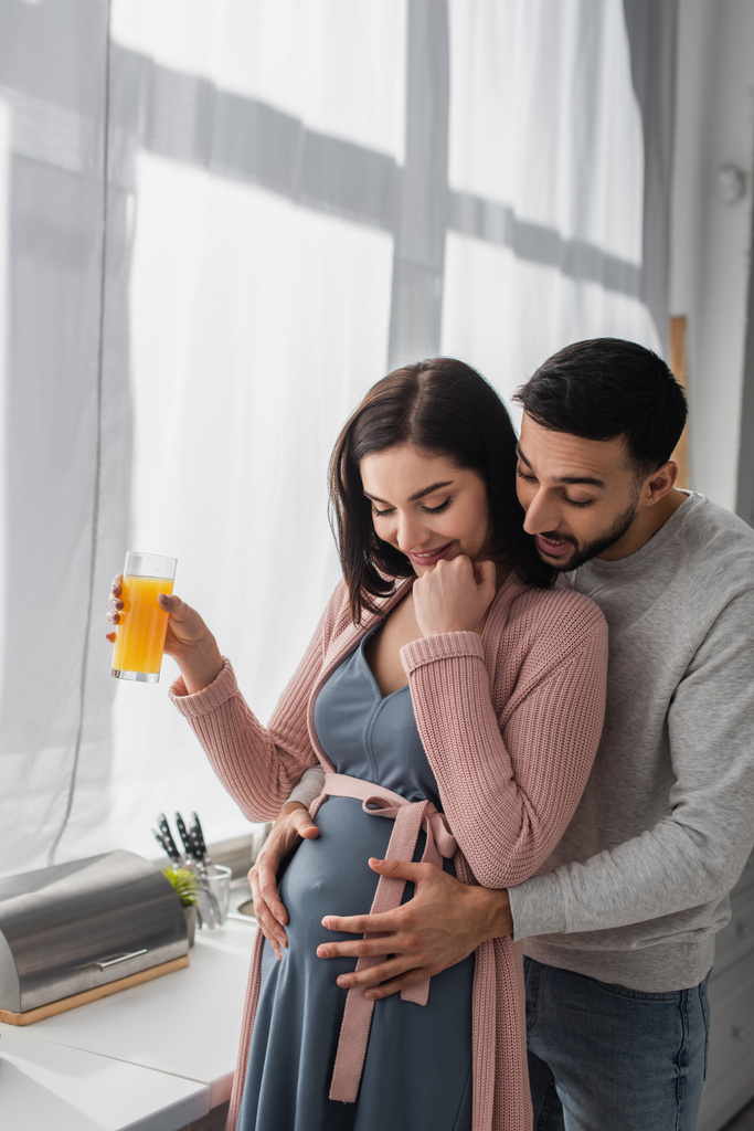 excité jeune homme embrassant doucement femme enceinte avec du jus d'orange dans la cuisine - Photo, image