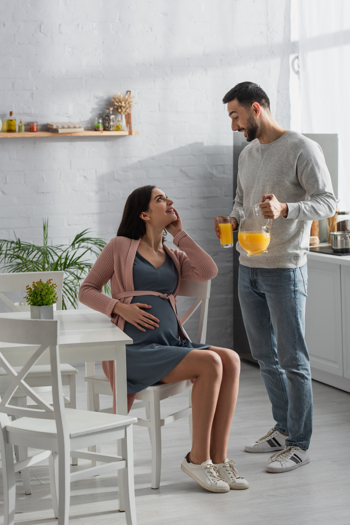 jovem sorridente de pé com frasco e copo de suco de laranja perto de mulher grávida na cozinha - Foto, Imagem