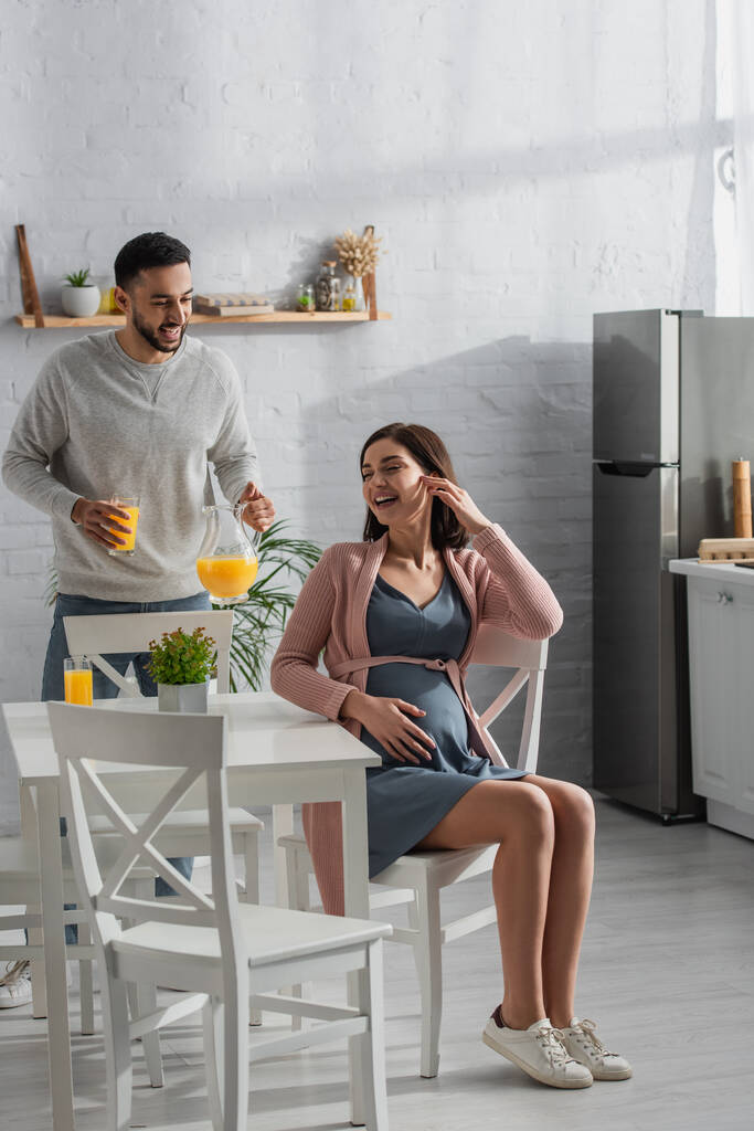 улыбающийся молодой человек, стоящий с банкой и стаканом апельсинового сока рядом с беременной женщиной на кухне - Фото, изображение
