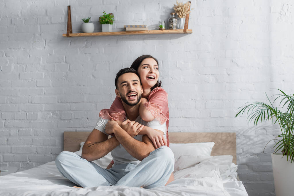 Lächelndes junges Paar sitzt mit weißer Bettwäsche auf dem Bett und umarmt sich im Schlafzimmer - Foto, Bild
