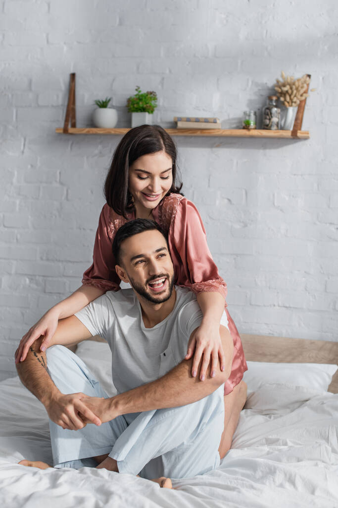 happy young couple sitting on bed with white linen and hugging in bedroom - Photo, Image