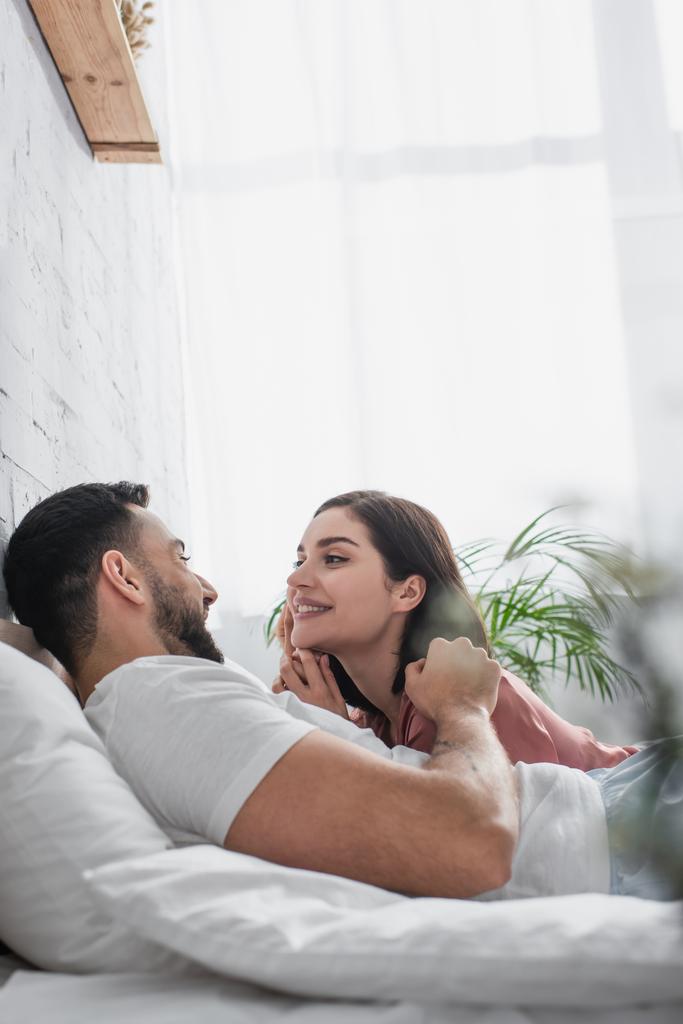 Mutlu genç adam beyaz çarşaflarla yatakta uzanıyor ve yatak odasında kız arkadaşının yüzüne nazikçe dokunuyor. - Fotoğraf, Görsel