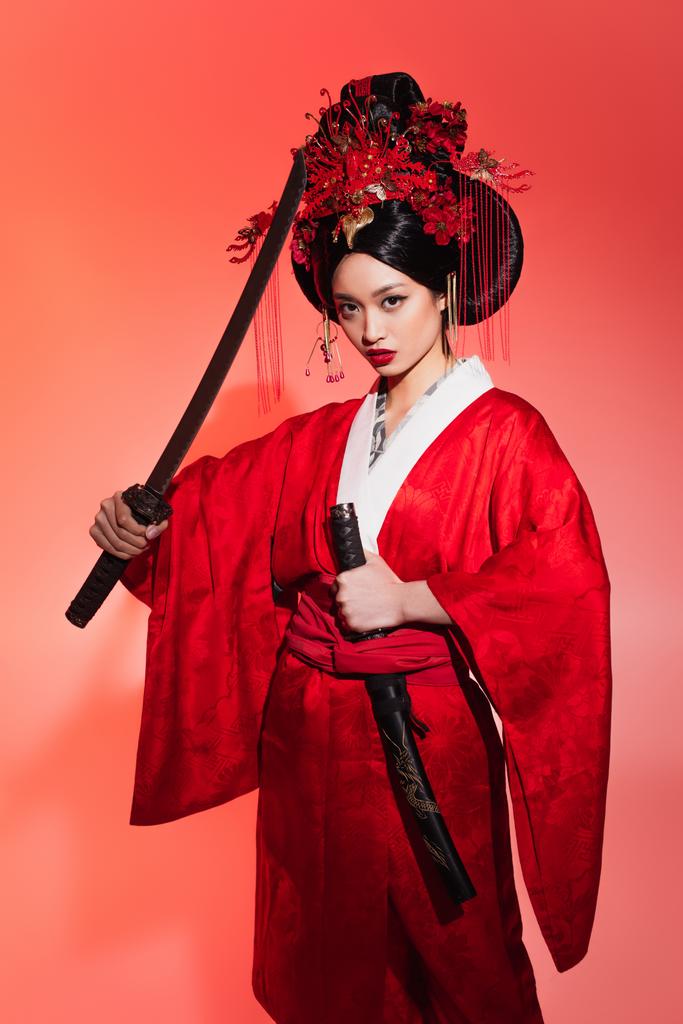 Νεαρή Ασιάτισσα με κιμονό κρατώντας σπαθί και θήκη σε κόκκινο φόντο  - Φωτογραφία, εικόνα