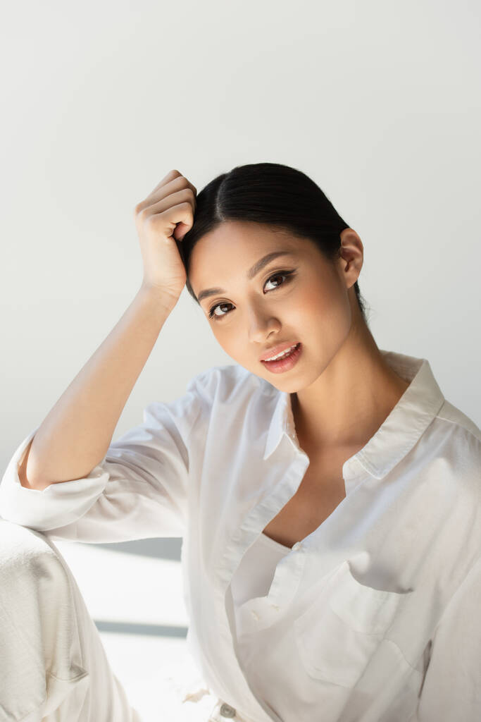 Pozitivní japonská žena v bílém oblečení při pohledu na kameru na šedém pozadí  - Fotografie, Obrázek