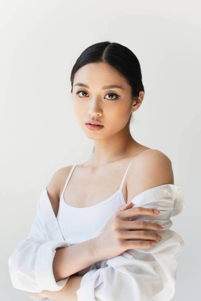 Jovem mulher asiática em roupas brancas tocando braço isolado em cinza - Foto, Imagem