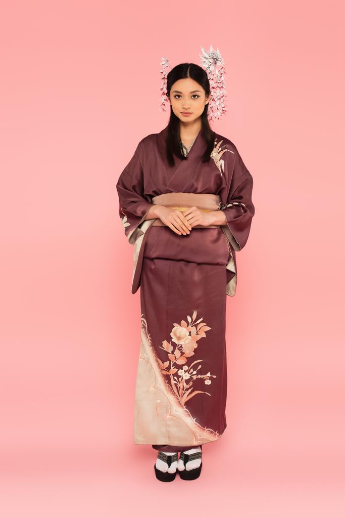 Longitud completa de mujer asiática en kimono y chanclas mirando a la cámara sobre fondo rosa - Foto, Imagen